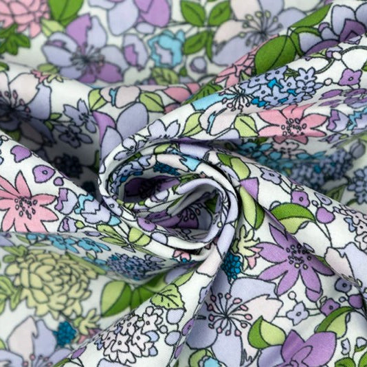 A close-up of a floral printed fabric with a white base and a pattern of purple, pink, blue, and green flowers and leaves, arranged in soft folds.