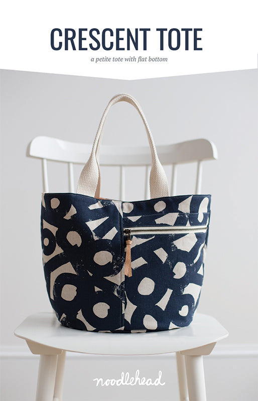 A blue and white print crescent tote bag sits on a white chair against a white background.
