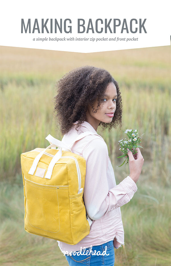 A person with curly brown hair is wearing a yellow backpack in a field while holding wildflowers.