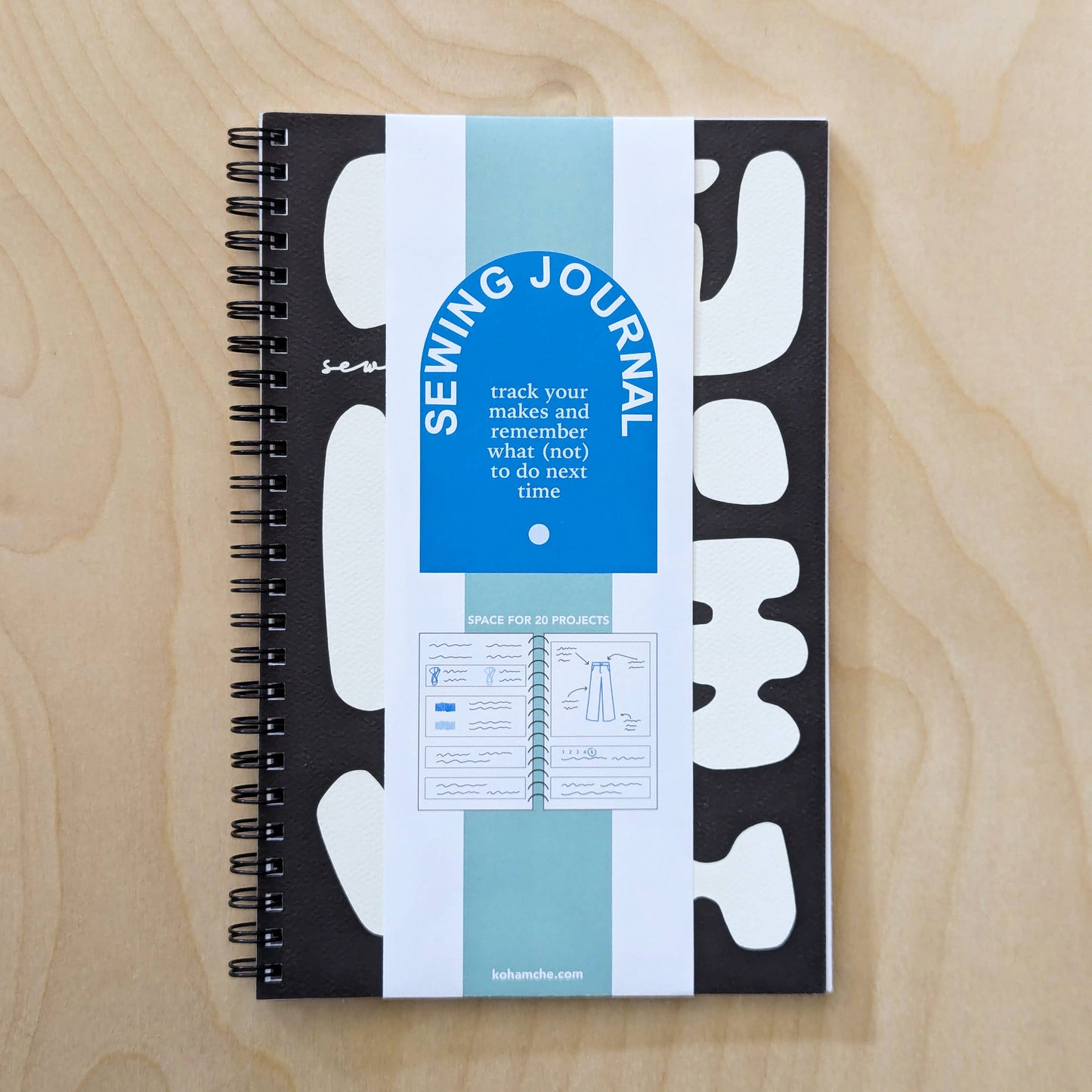 A black and white spiral bound notebook with a blue tag labeled “Sewing Journal” sits on a light wooden table.