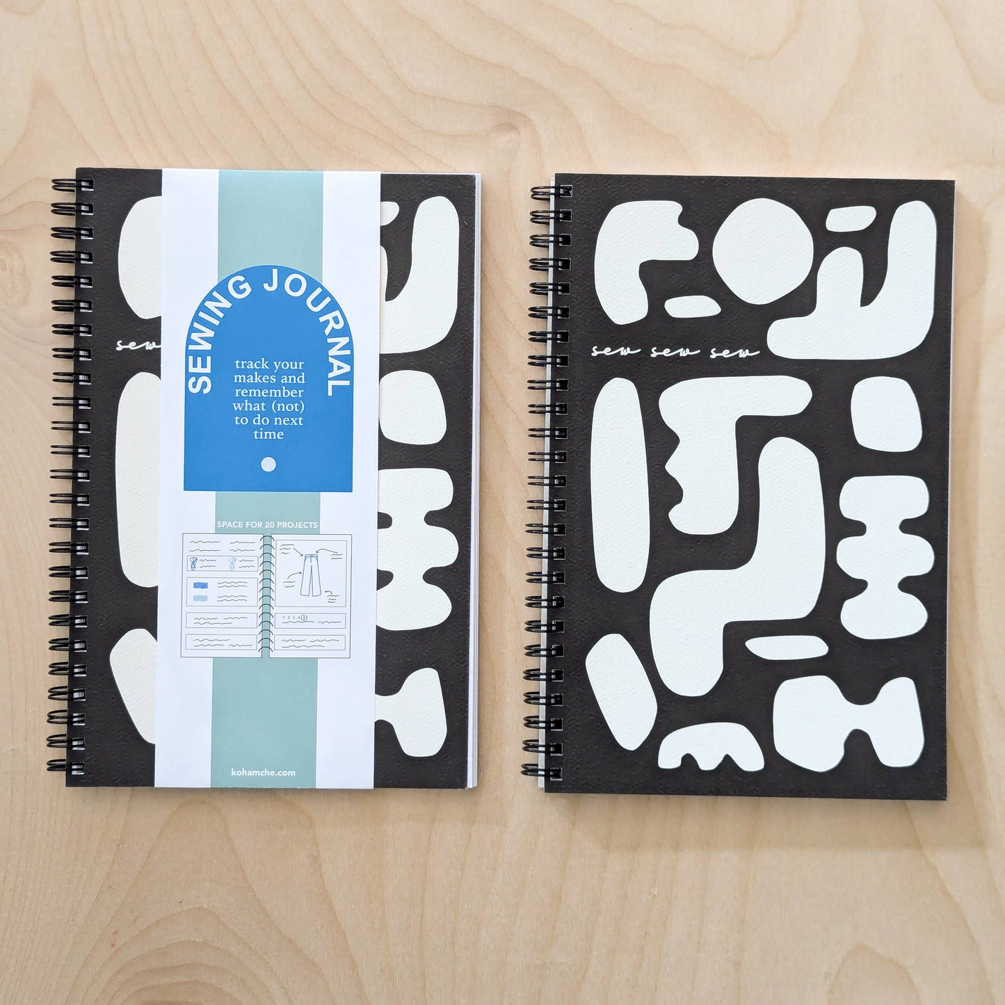 Two black and white spiral bound notebooks - one with a blue tag labeled “Sewing Journal”  - sit on a light wooden table.