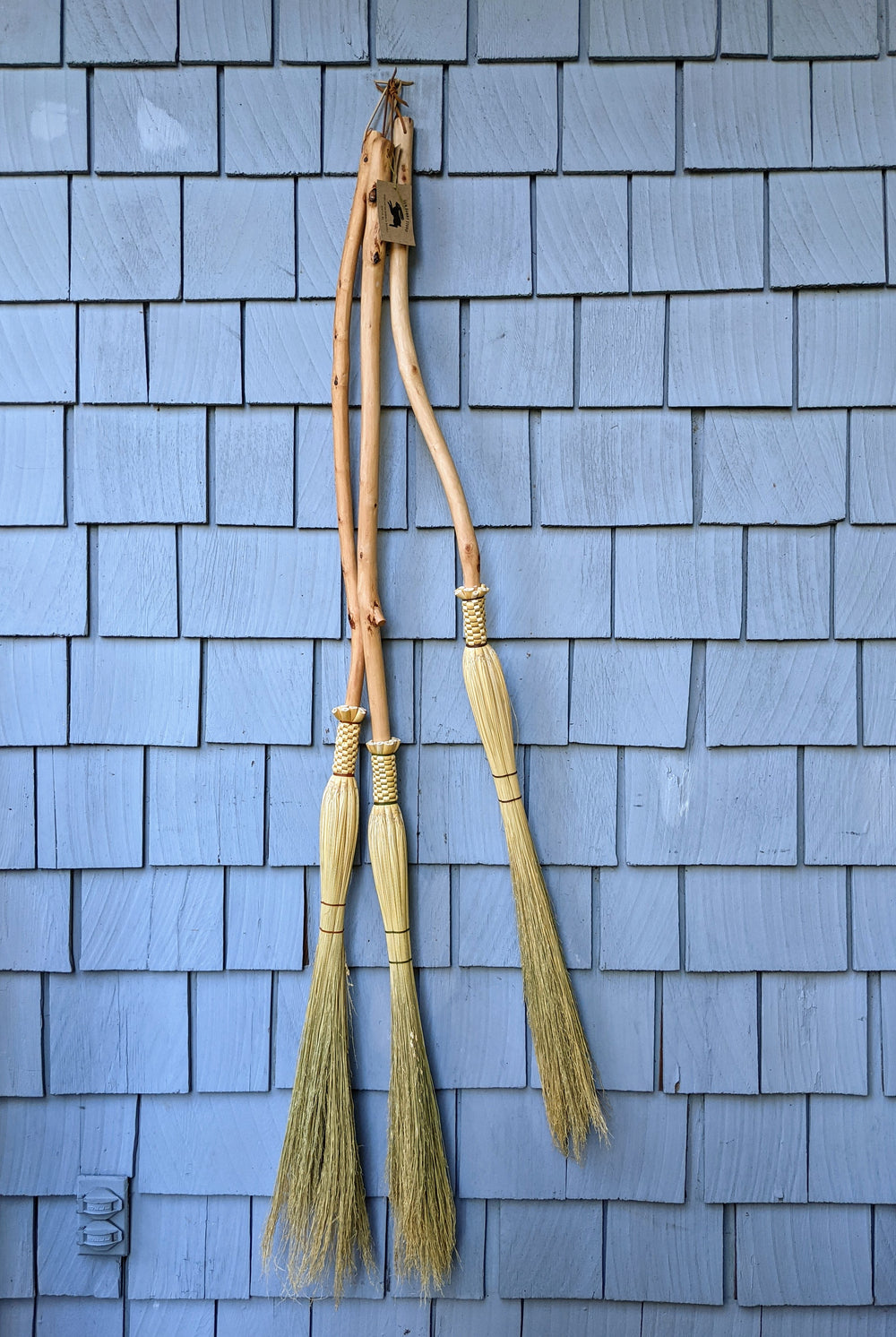 Three cobwebbers (long skinny hand brooms) hanging from a hook on an outdoor wall made in a crafting class at Wyldwood Creative Renton.