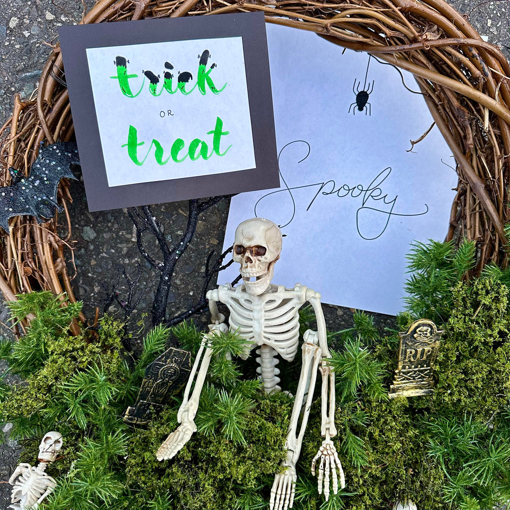 Frankenstein style hand lettering sign that says "trick or treat" and a curly font that said "spooky" with a spider - both on display with a Halloween scene of skeletons and tombstones - made in a Spooky hand letter class at Wyldwood Creative in Renton